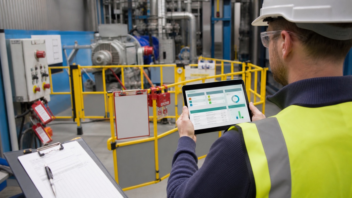 A site supervisor viewing a live permit-to-work status board on a tablet beside a controlled maintenance area with barriers, isolation tags and contractor sign-in evidence.