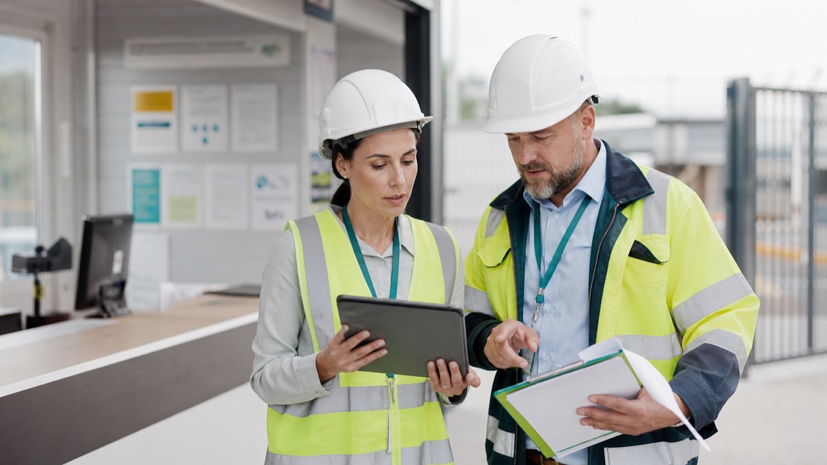 A facilities manager and contractor supervisor reviewing a permit, RAMS record and live action dashboard together at a bright industrial site entrance.